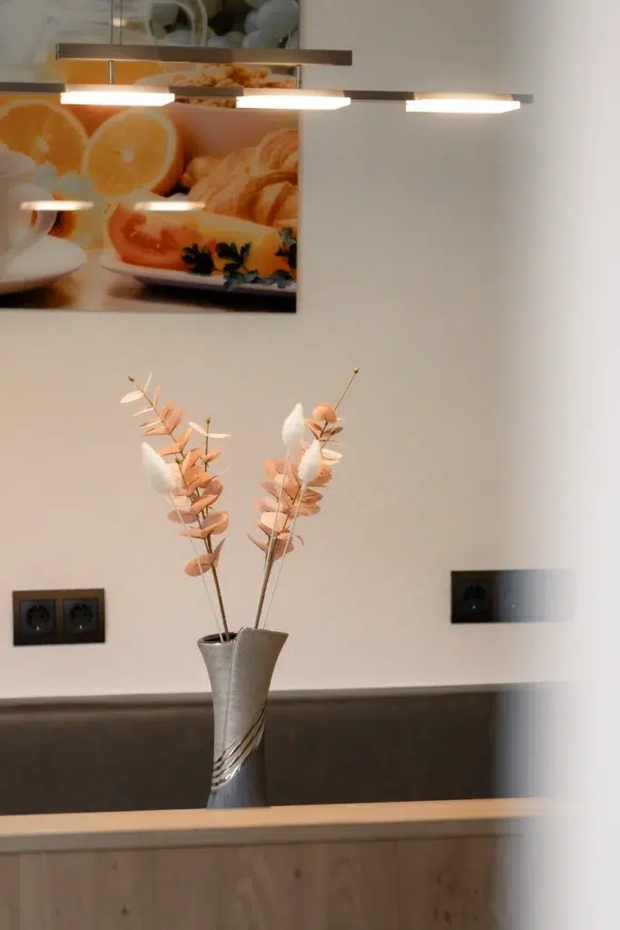 Eine minimalistische beige Vase hält getrocknete orangefarbene und weiße Blumen auf einem Holztisch. Darüber hängt eine moderne Leuchte und man sieht eine Küchentheke. Im Hintergrund ist ein Wandbild mit Orangen und Brot teilweise zu sehen.