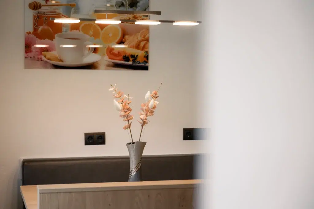 Auf einem Holztisch steht eine Vase mit pfirsichfarbenen dekorativen Zweigen. Im Hintergrund ist an einer Wand ein Foto einer Frühstücksszene mit Orangen und einer Teetasse zu sehen. Darüber hängt eine moderne Beleuchtung, die die Szene erhellt.