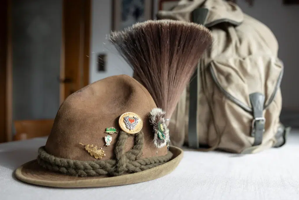 Auf einem Tisch liegt ein mit Nadeln und einer großen Feder geschmückter Alpenhut. Im Hintergrund ist ein leicht unscharfer, beiger Rucksack mit grünen Riemen zu sehen.