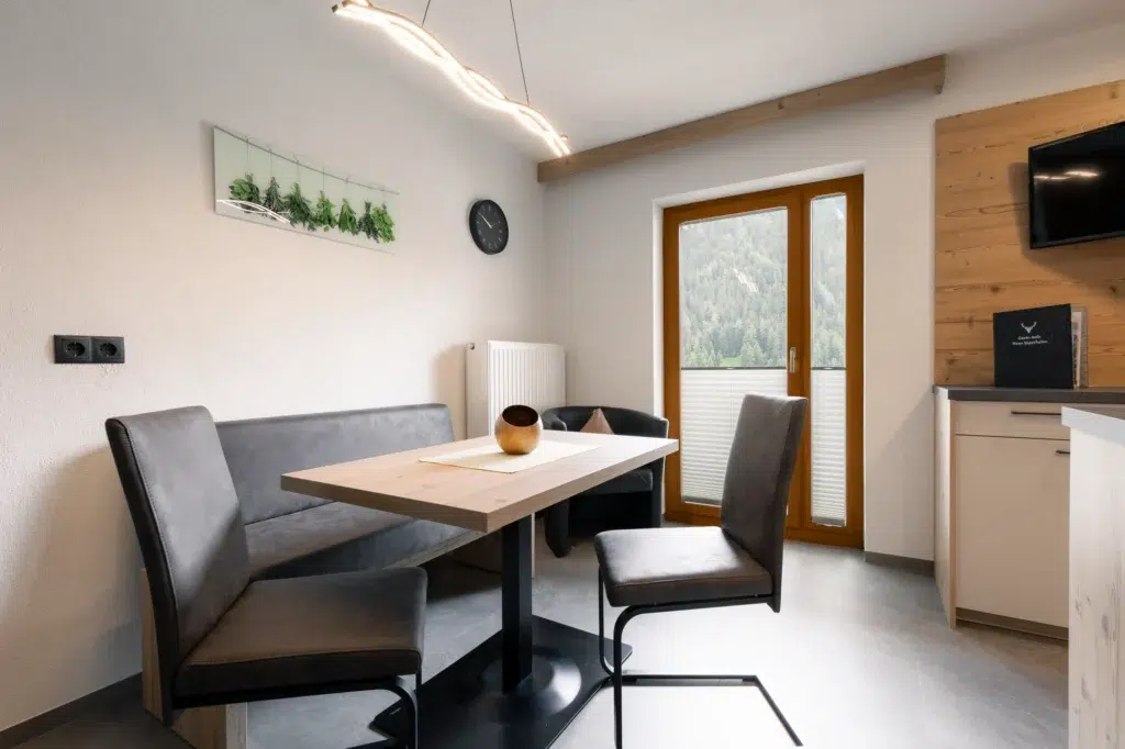 Moderner Essbereich mit einem Holztisch und dunklen Stühlen, einer Bank auf einer Seite, einem dekorativen Holzwandpaneel und einem an der Wand montierten Fernseher. Über dem Tisch hängt eine einzigartige Leuchte. Ein großes Fenster bietet einen malerischen Blick auf die Berge draußen.
