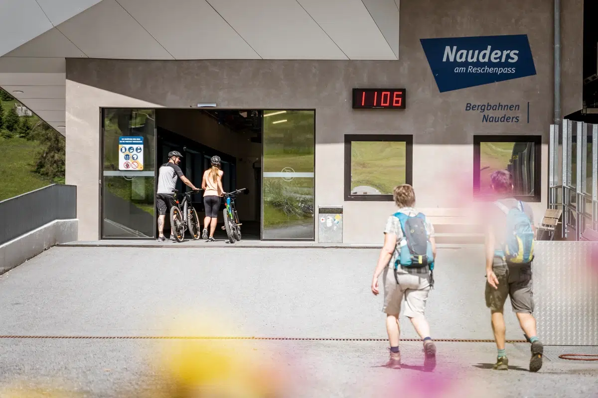 Zwei Radfahrer betreten mit ihren Rädern eine Seilbahnstation, während zwei Wanderer mit Rucksäcken auf den Eingang zulaufen. Auf der Station sind die Aufschriften „Nauders am Reschenpass“ und „Bergbahnen Nauders“ zu sehen. Eine Digitaluhr zeigt 11:06 Uhr. Im Vordergrund sind rosa Blumen zu sehen.