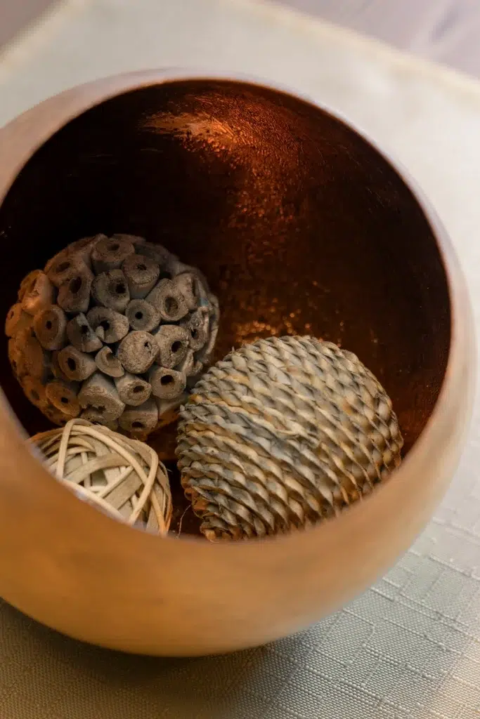 Eine Holzschale mit drei dekorativen Objekten: einem Tannenzapfen, einem geflochtenen Ball und einer Samenkapsel. Die Innenseite der Schale hat eine warme, kupferähnliche Oberfläche und steht auf einer strukturierten, hellen Stoffoberfläche.
