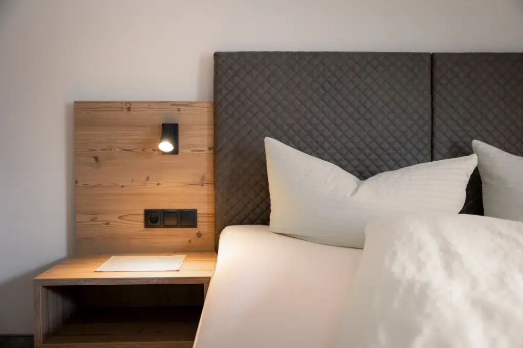 Neben einem Nachttisch aus Holz steht ein ordentlich gemachtes Bett mit weißer Bettwäsche und einem gepolsterten Kopfteil. An der Holzplatte ist über zwei Steckdosen eine kleine Leselampe angebracht.