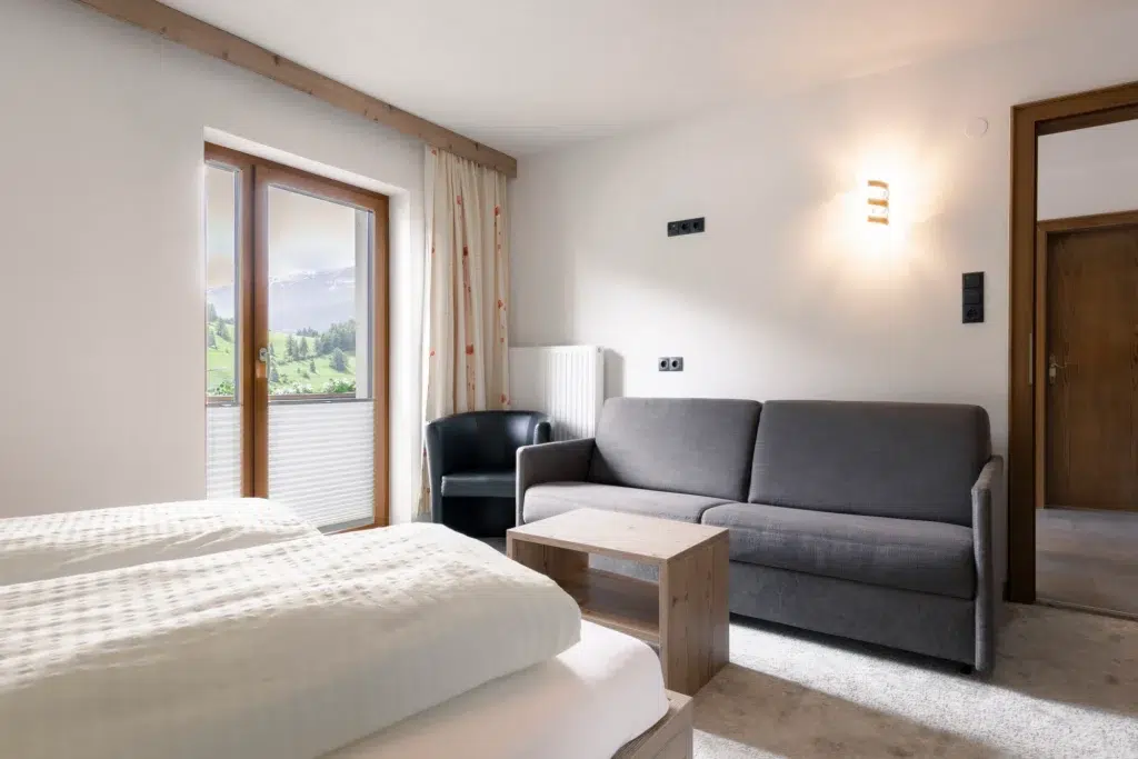 Ein gemütliches Hotelzimmer mit einem Bett mit weißer Bettwäsche, einem grauen Sofa, einem kleinen Holztisch und einem schwarzen Sessel. Ein Fenster mit teilweise zugezogenen Vorhängen bietet den Blick auf eine grüne Landschaft und Berge. Warmes Licht trägt zur einladenden Atmosphäre bei.