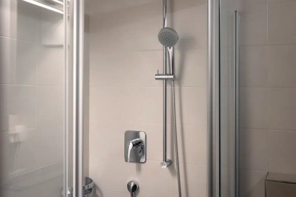 Moderne Dusche mit Glastüren, einem eleganten silbernen Duschkopf auf einer verstellbaren Stange, einem quadratischen, an der Wand montierten Wasserhahn und einem glatten, hellen Fliesenhintergrund.