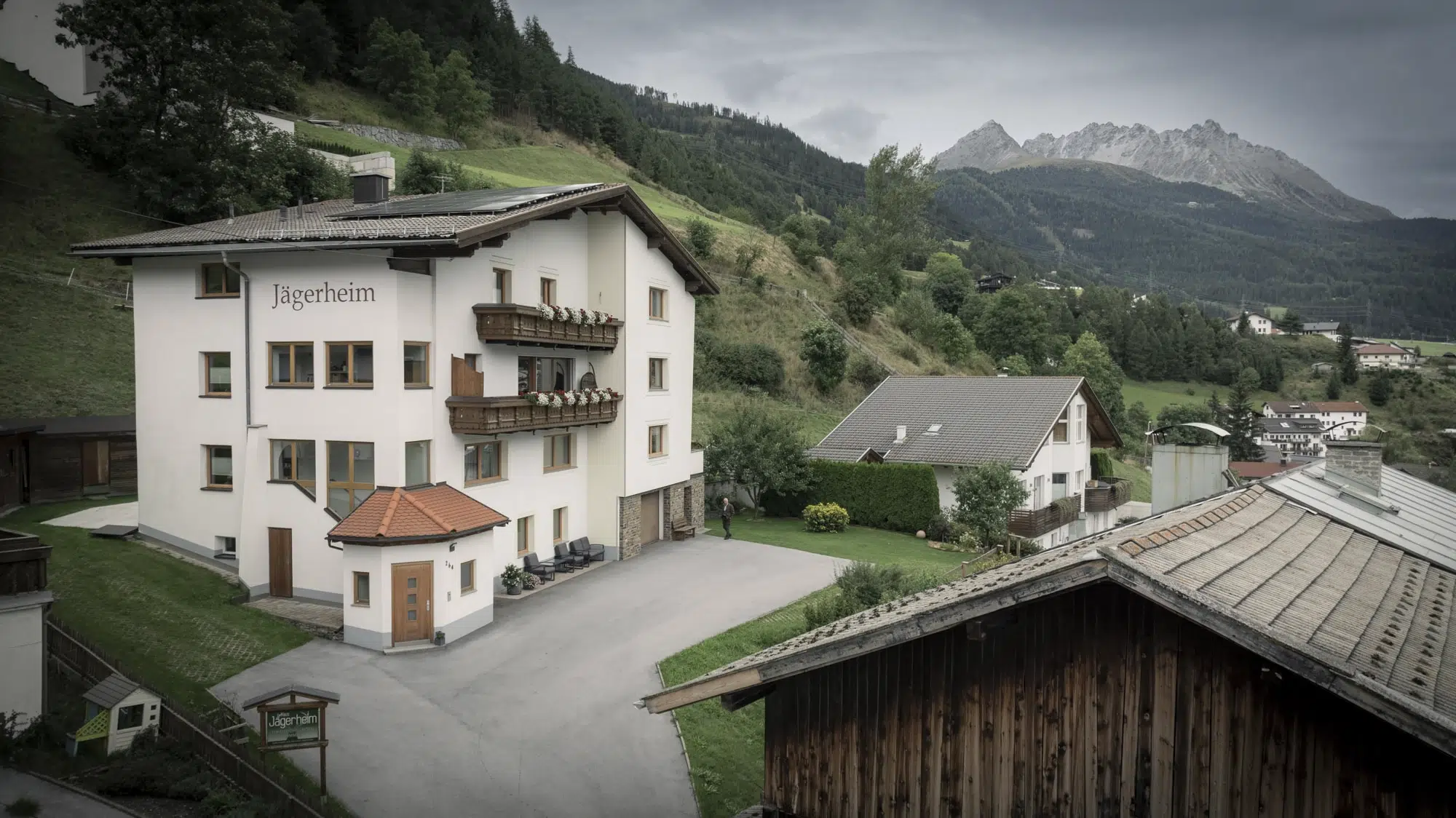 Ein großes Alpenhaus mit Balkonen, beschriftet mit „Jägerheim“, liegt eingebettet in einer Berglandschaft. Die Szene umfasst nahe gelegene Häuser, grüne Hügel und ferne Berge unter einem bewölkten Himmel.